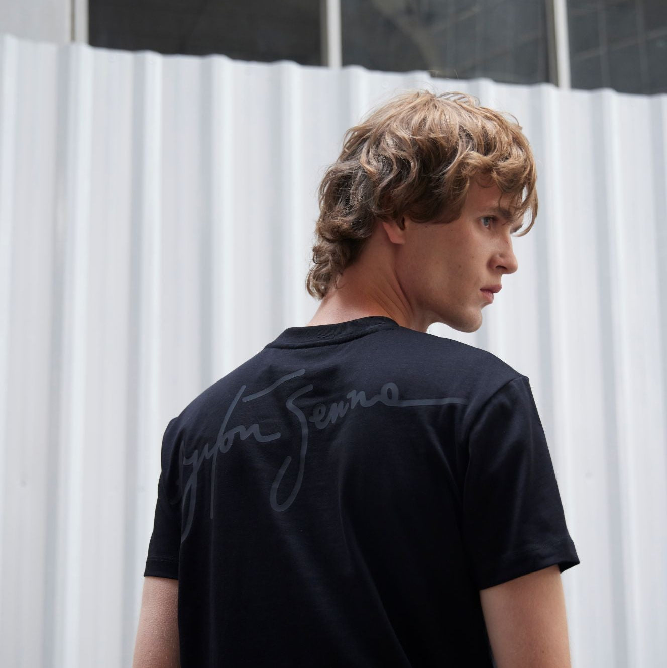 Person wearing a black t-shirt with a brand logo, standing against a white curtain.