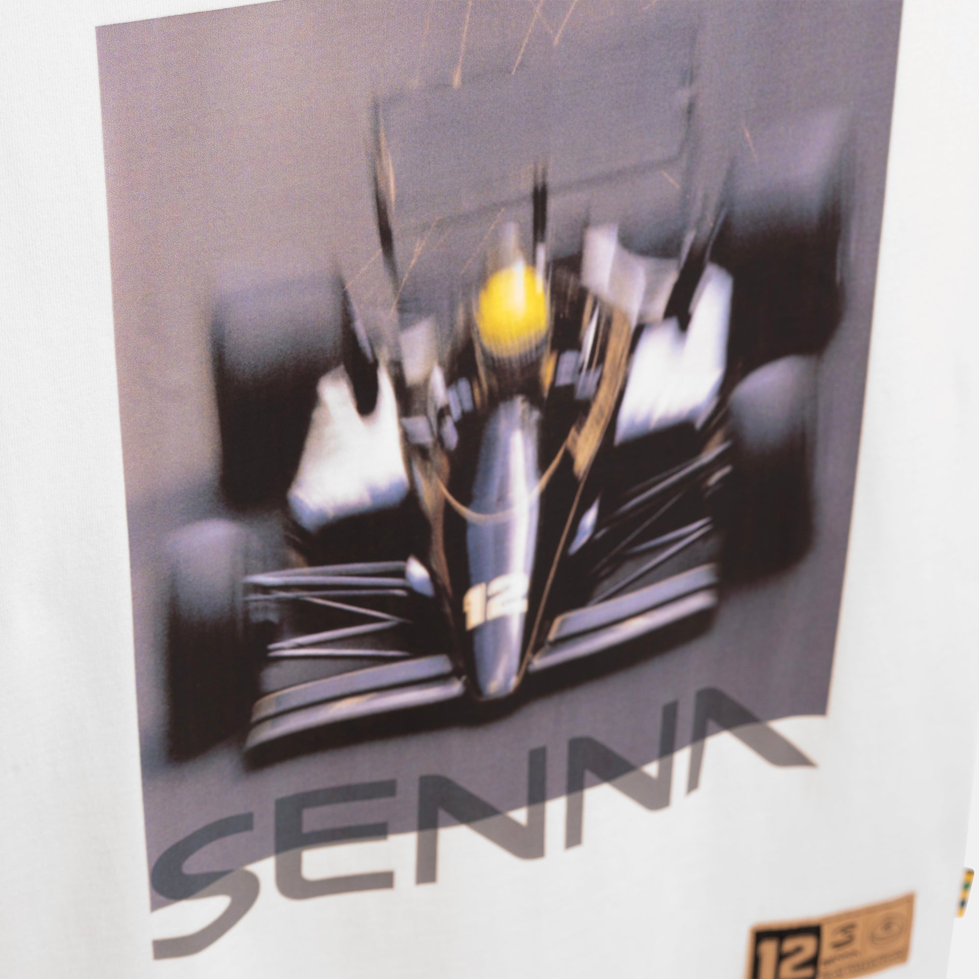 A close-up of a cream colored tee-shirt featuring a printed graphic of a black Formula 1 race car with a yellow helmet and the name 'SENNA' in large gray letters below. A small gold '12' sticker is visible in the bottom right corner.