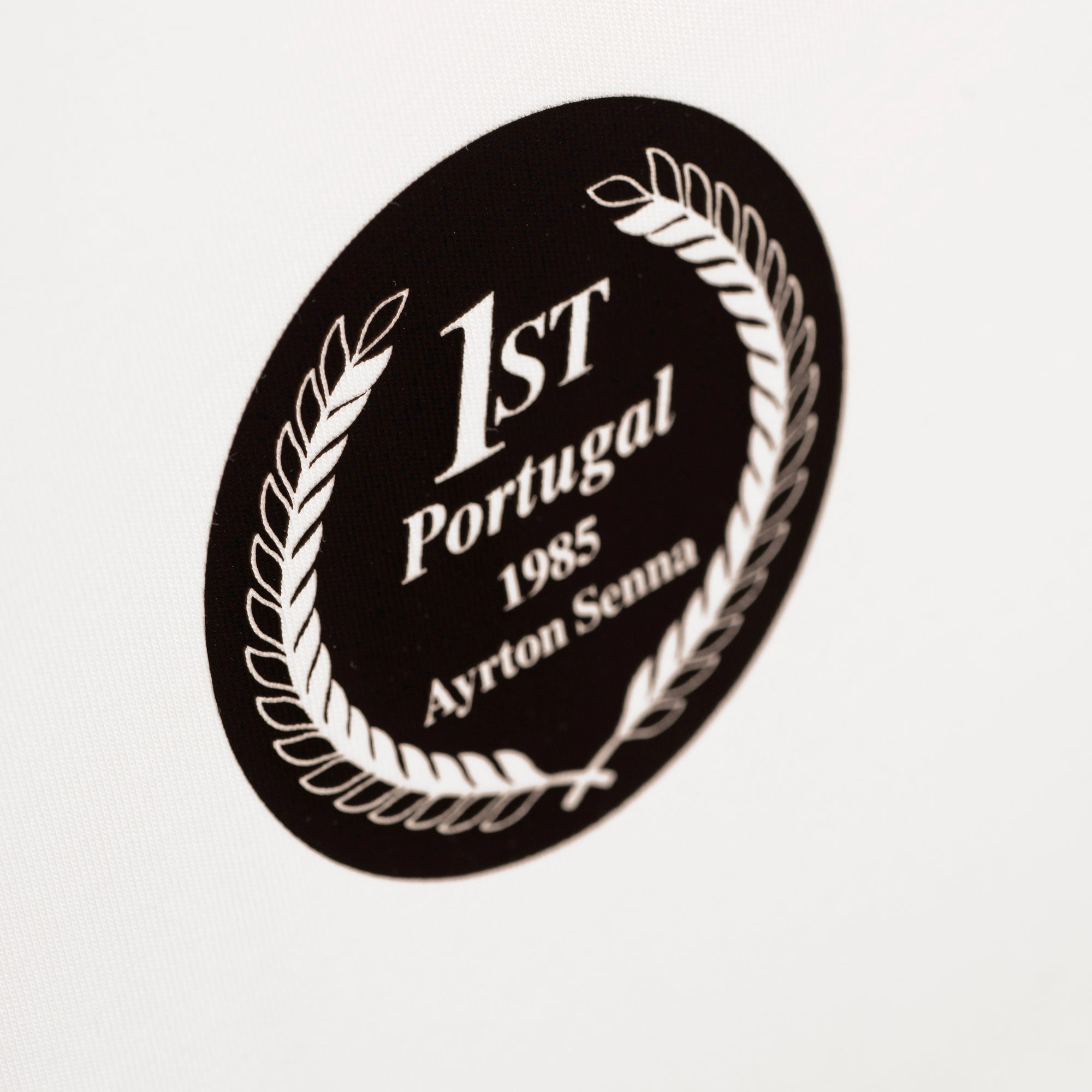 A t-shirt close-up of a circular black wreath emblem with white text reading '1st Portugal 1985 Ayrton Senna'.