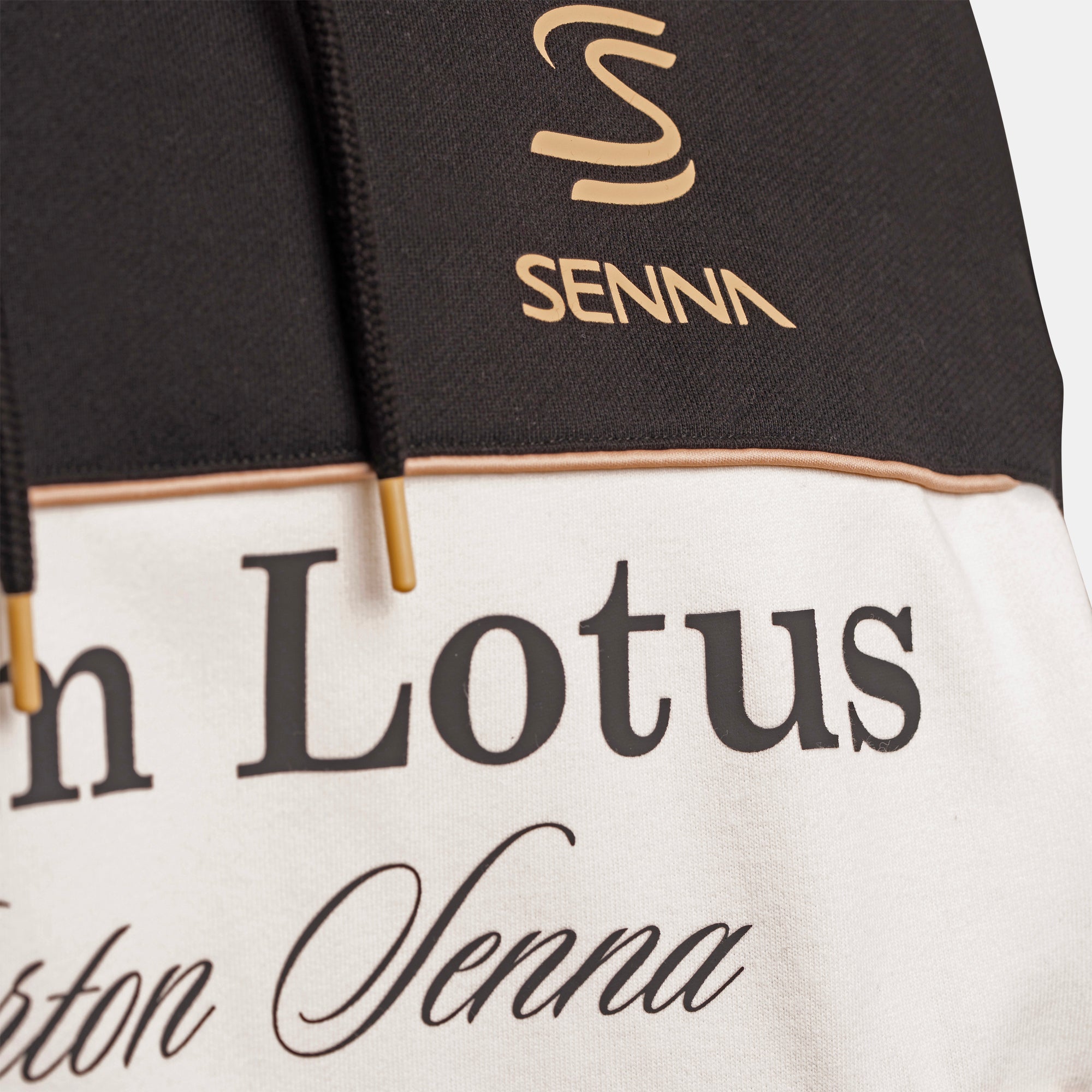 Close-up of a colorblock jacket featuring a black upper section with a gold Senna logo, a cream lower section with black text Team Lotus, and gold ended drawstrings.