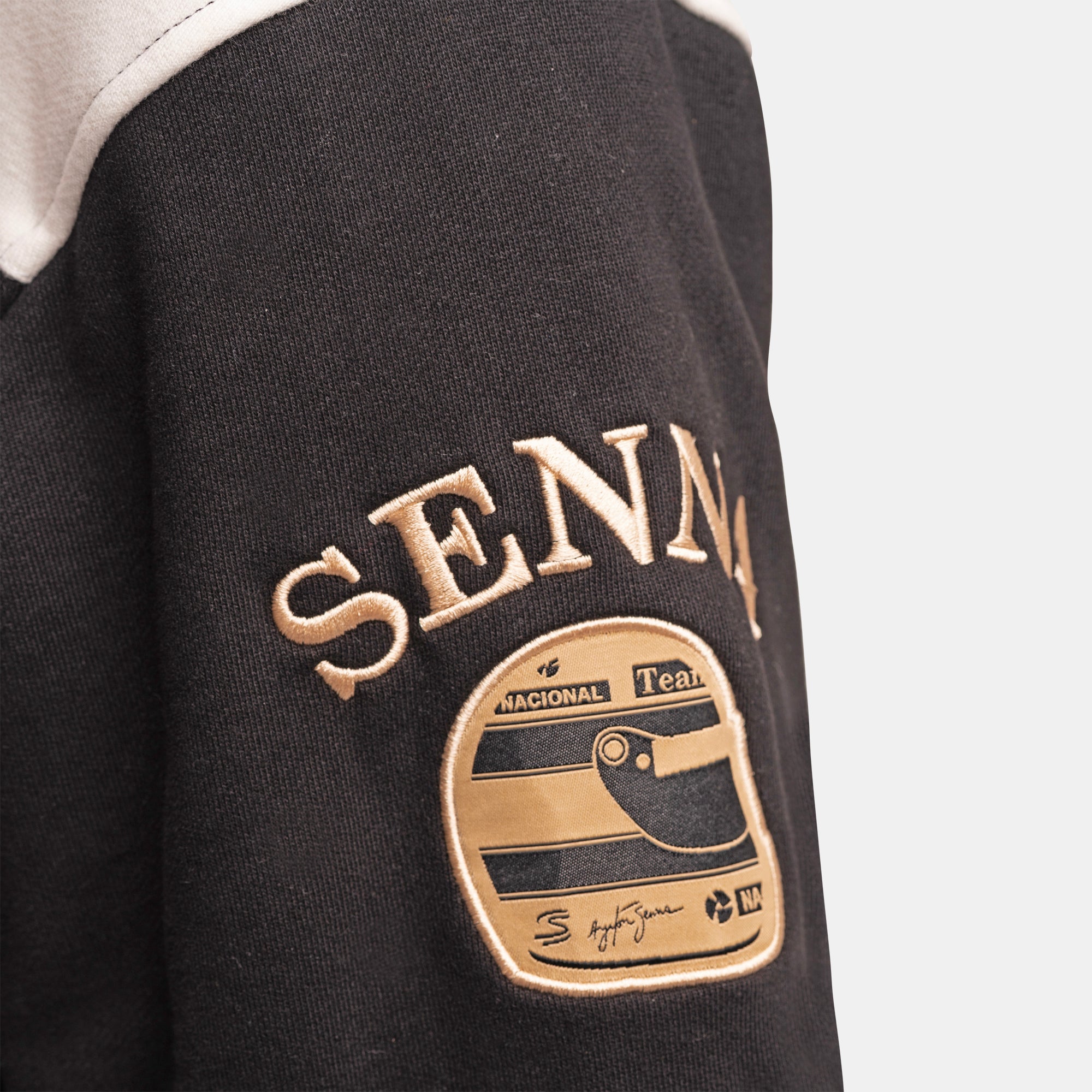 Close-up of a black varsity-style jacket featuring a gold embroidered patch with a helmet graphic and the word 'SENNA' above it, against a light gray background.