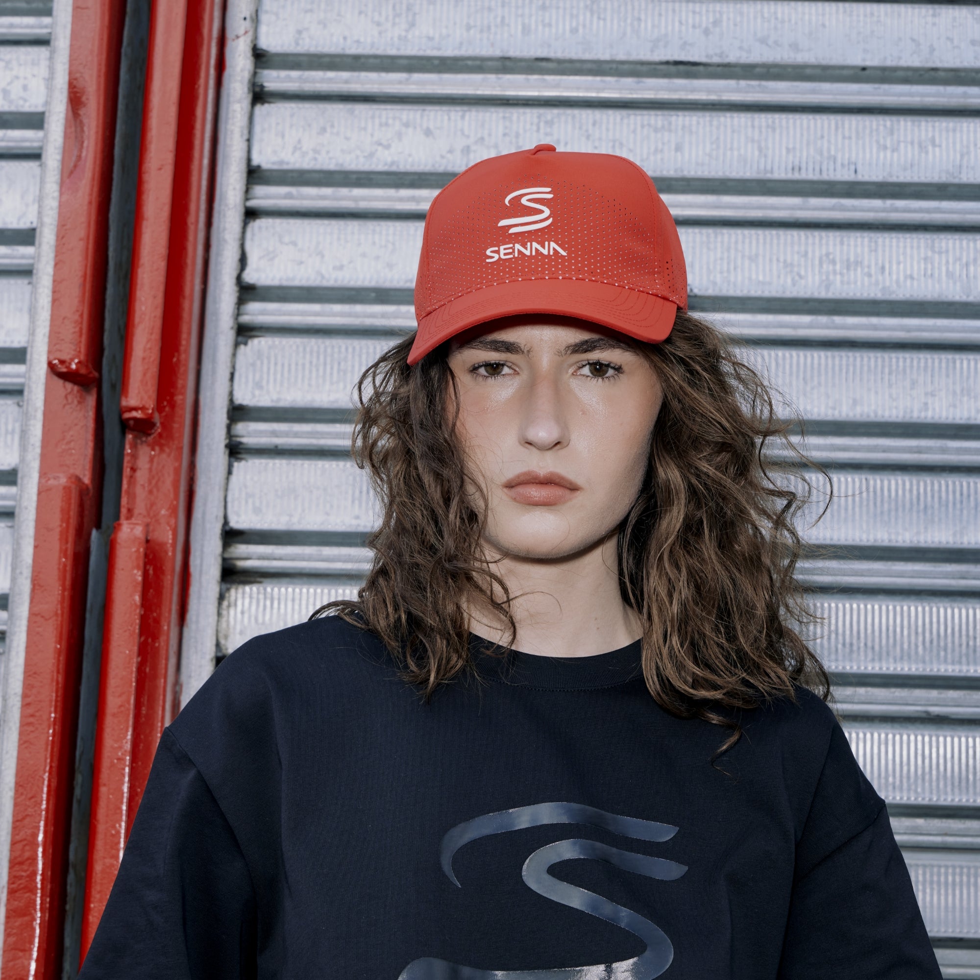 Person wearing a red Senna leisure cap and black t-shirt with a Senna logo, standing in front of a metallic shutter.