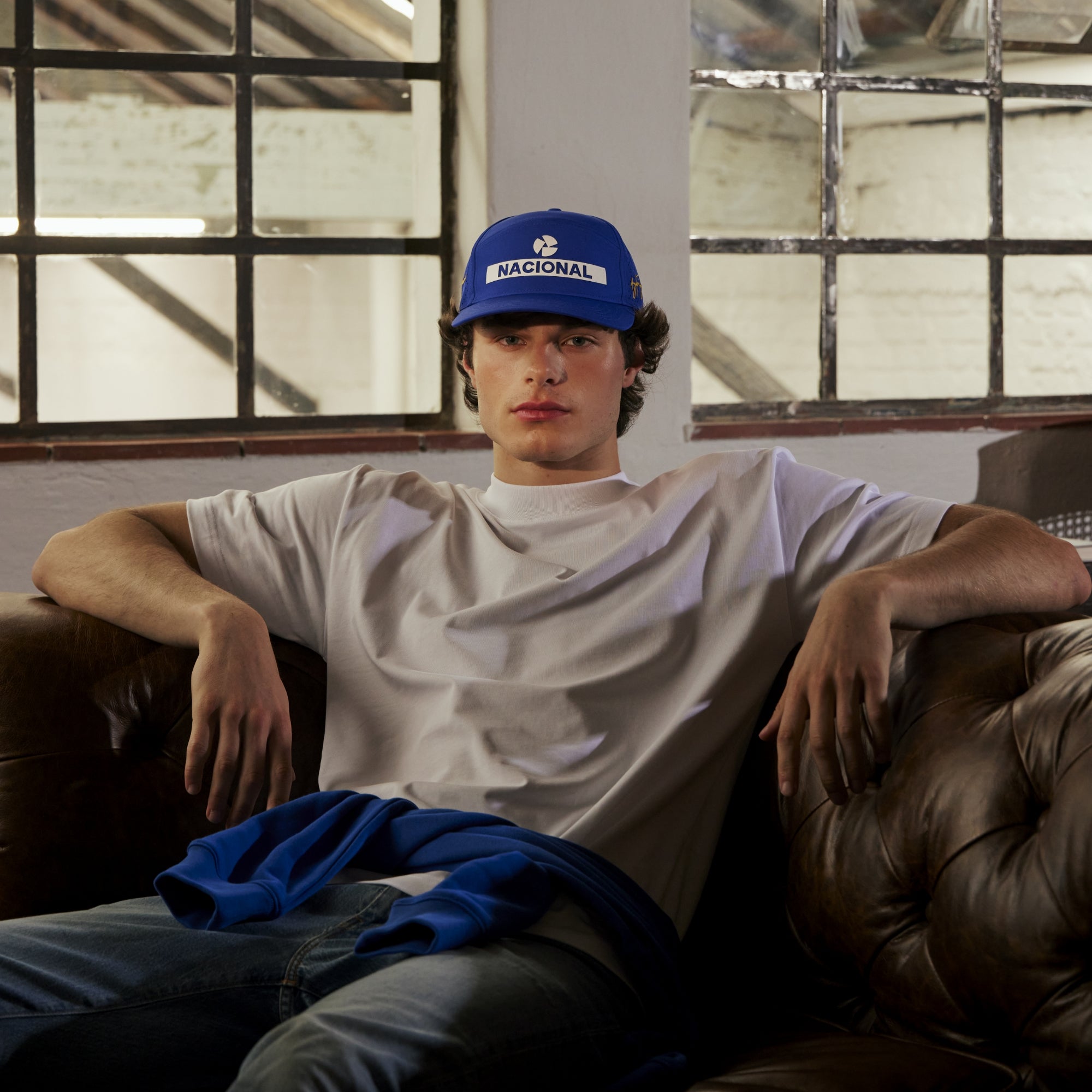 Person wearing a blue cap with 'Nacional' text, sitting on a brown leather couch in a room with large windows.