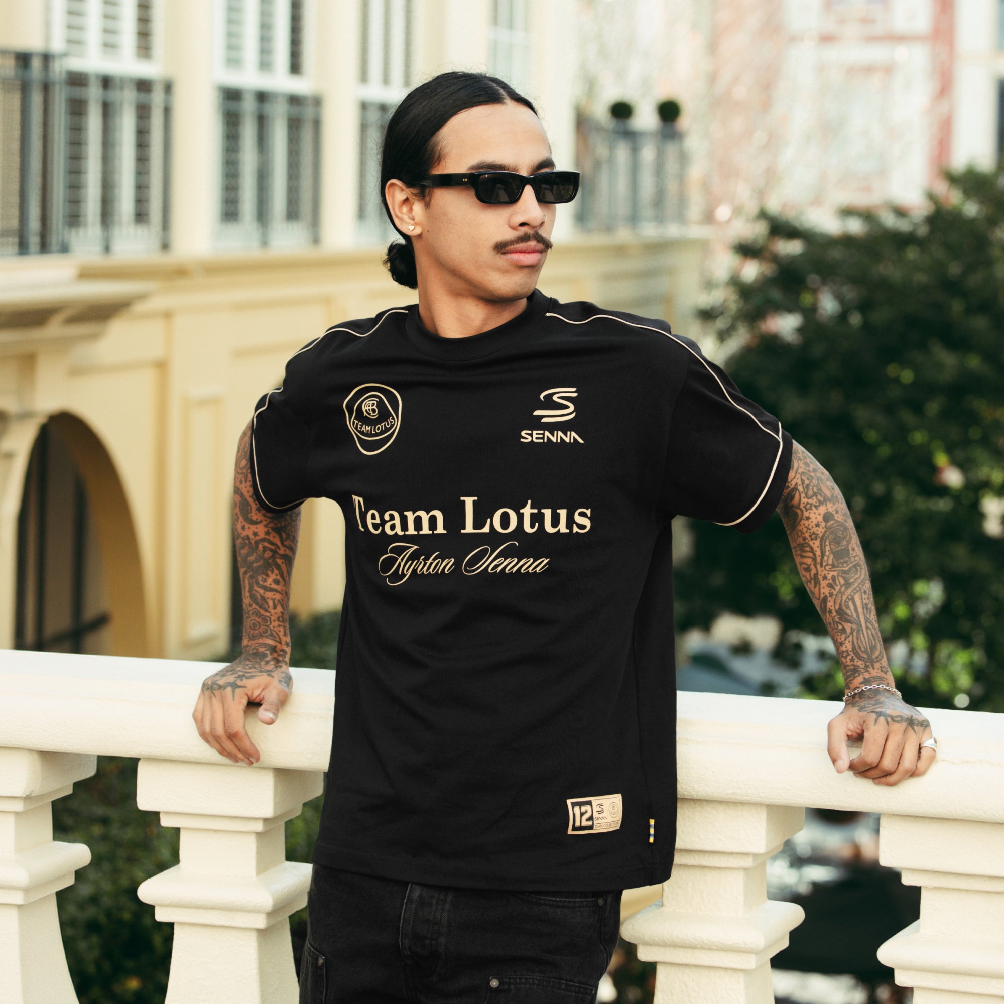 Man leaning against balcony wearing dark sun glasses and a black and gold t-shirt with Team Lotus and Senna branded patches.