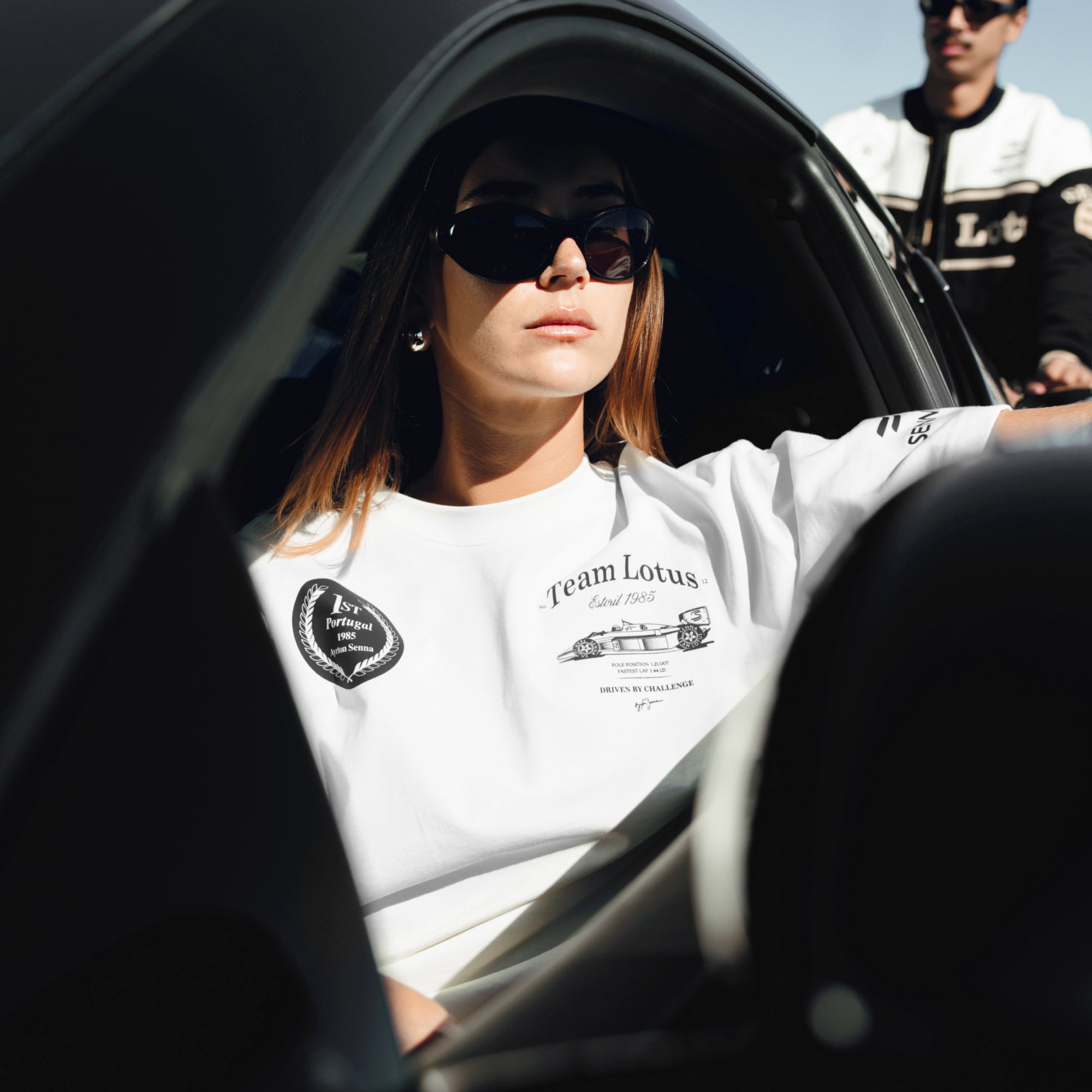 Woman lounging in car wearing cream colour oversize tee with Team Lotus and Senna graphics. Blurred man in background wearing black, white and gold jacket.