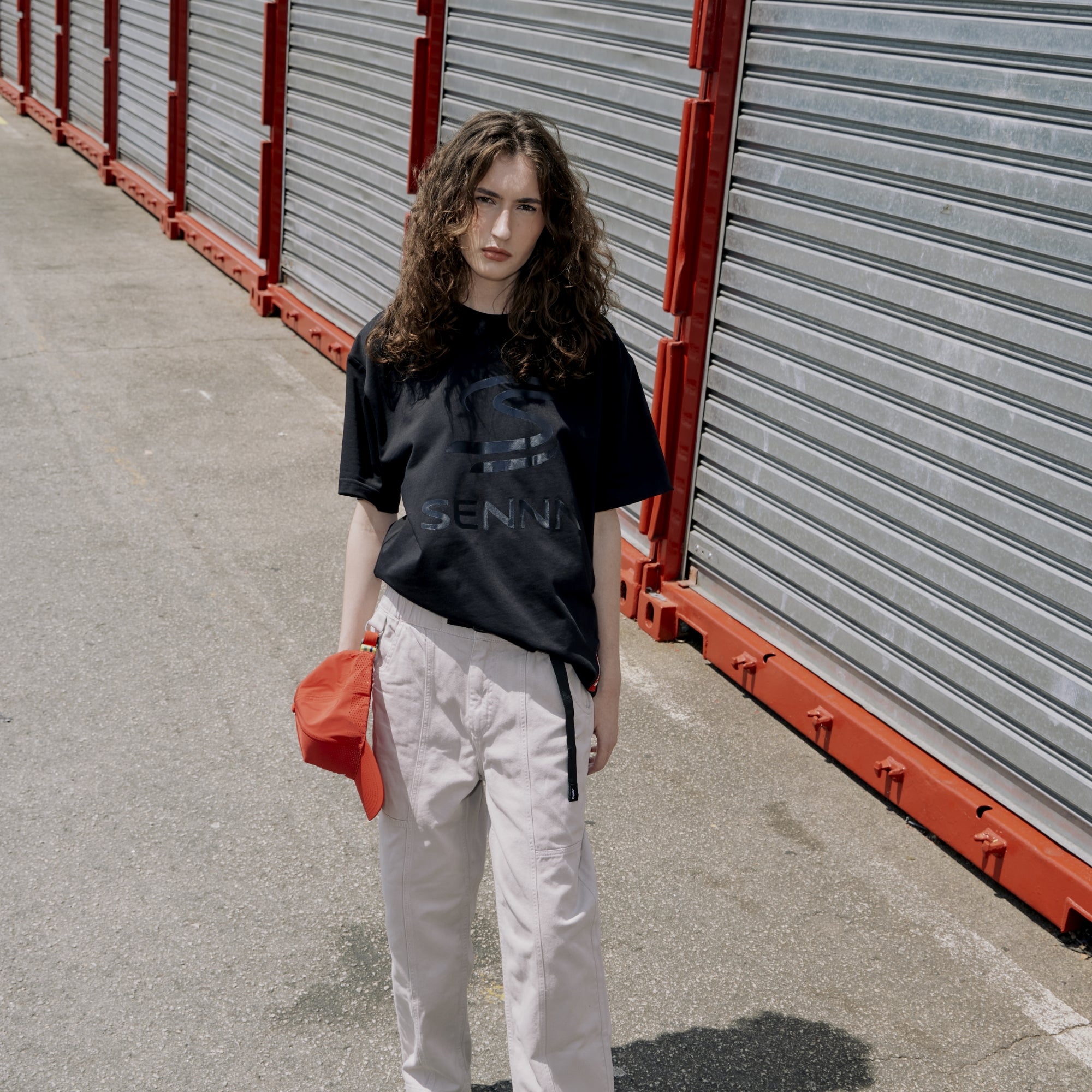 Person wearing a black t-shirt with text and light-colored pants, holding an red hat, standing in front of storage units.
