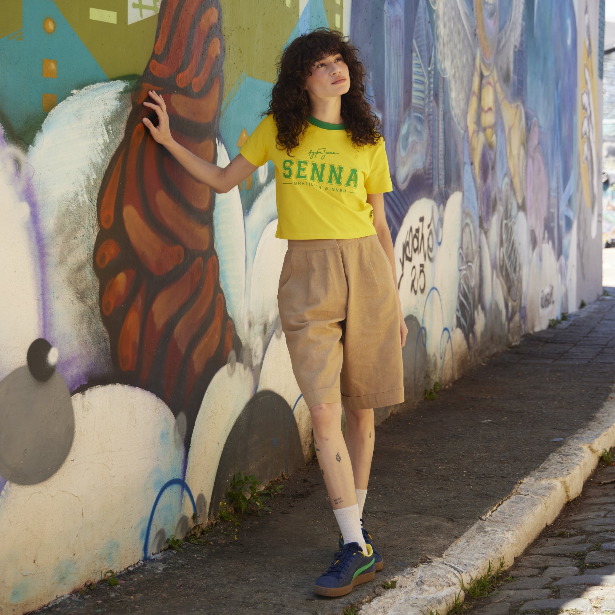 Person wearing a yellow shirt with 'SENNA' text, standing in front of a colorful mural.