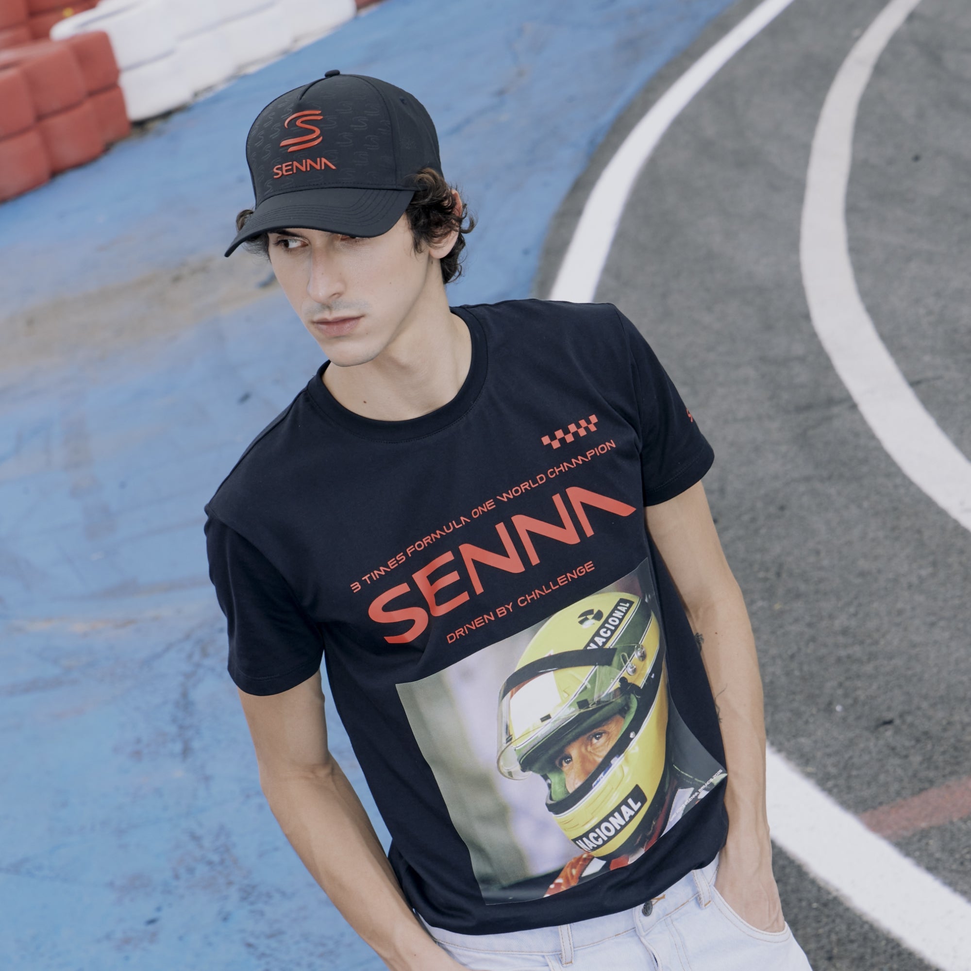 Person wearing a black t-shirt with 'Senna' branding and a helmet graphic, standing on a race track.