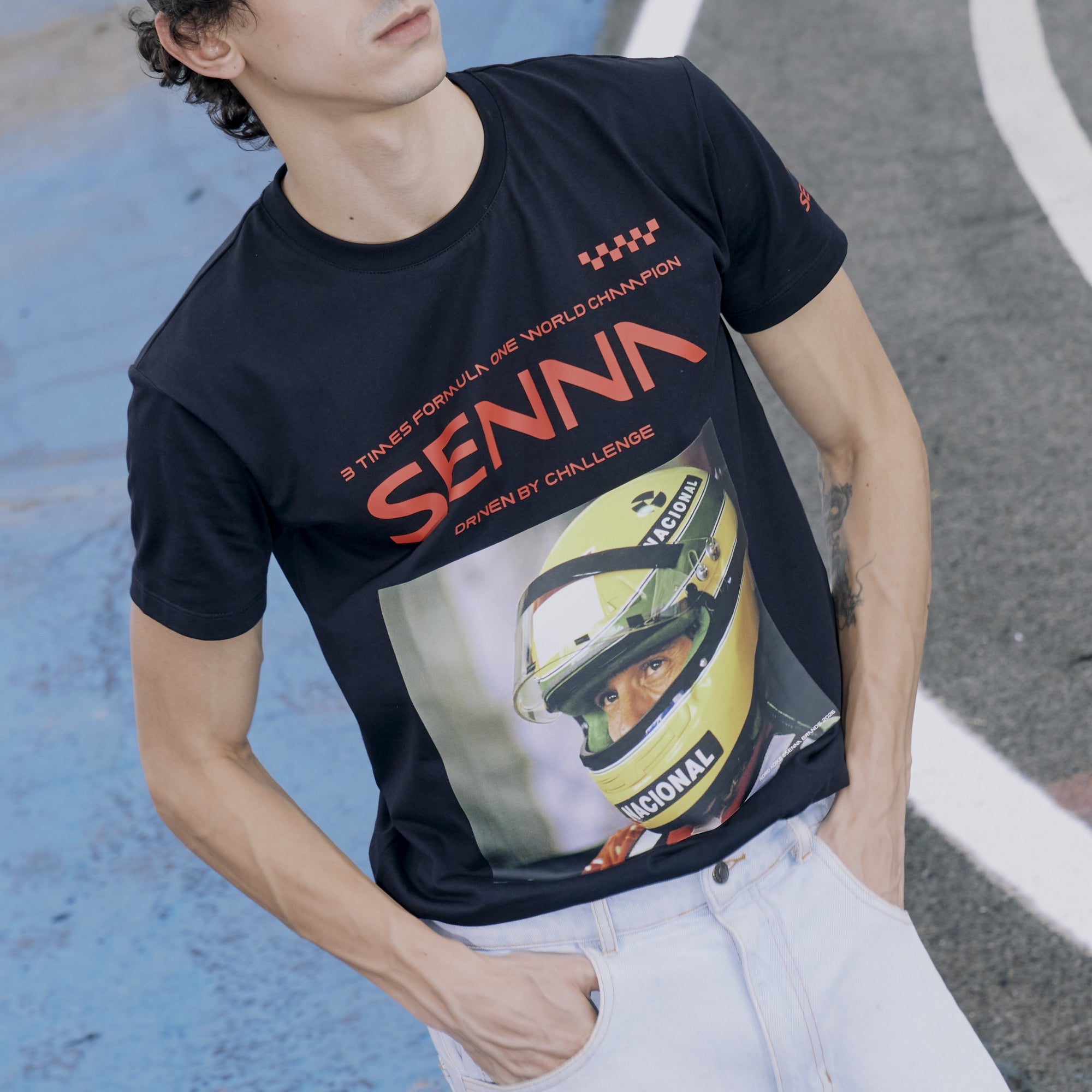Person wearing a black senna race collection driven by challenge t-shirt with a graphic and text, standing on a road.