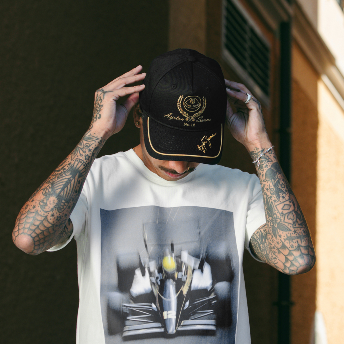 A tattooed man wearing a black and gold Team Lotus and Senna cap and a white graphic t-shirt with a race car print, posing outdoors.