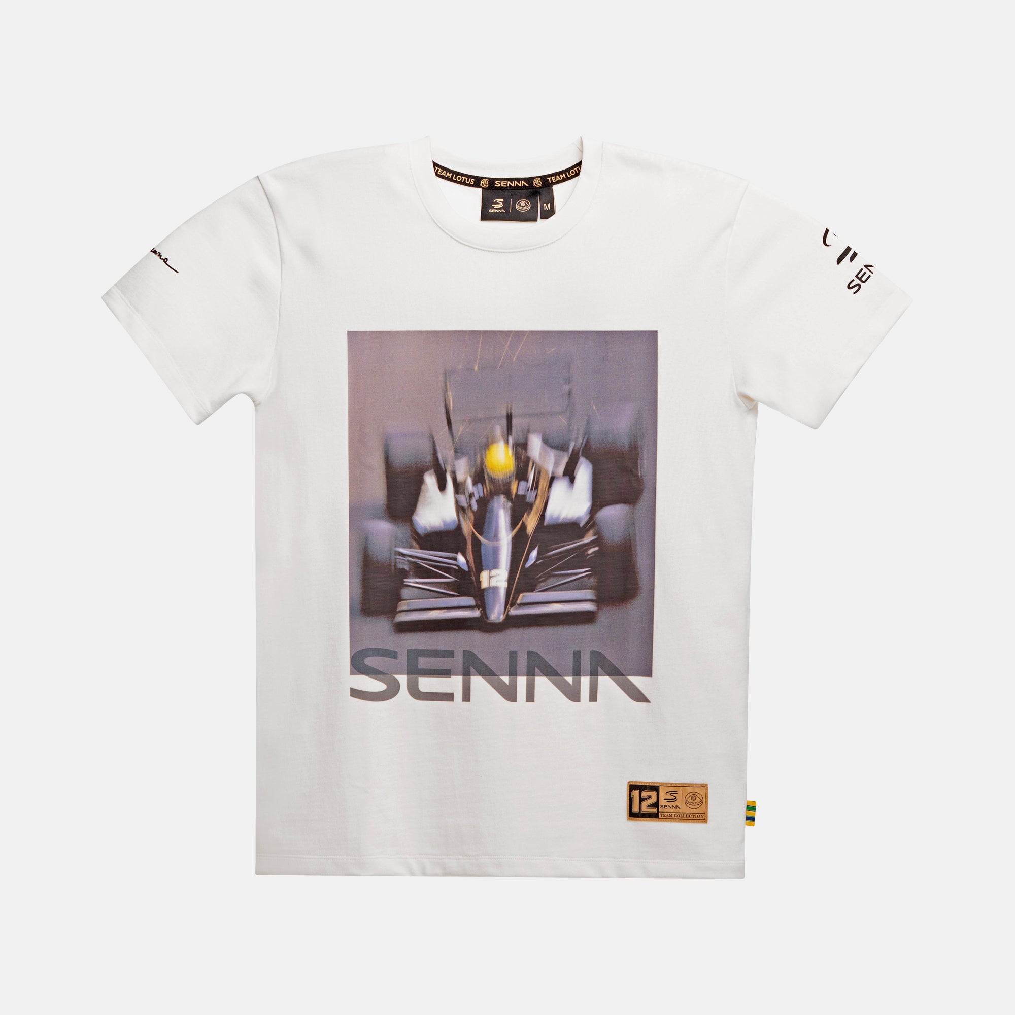 A cream colored short-sleeve t-shirt featuring a large graphic print of a Formula 1 race car on the front, with the brand name 'SENNA' printed below the graphic. The shirt also features small black graphics on the sleeves and a small gold brand label near the bottom right hem with '12' Senna X Team Lotus branding.