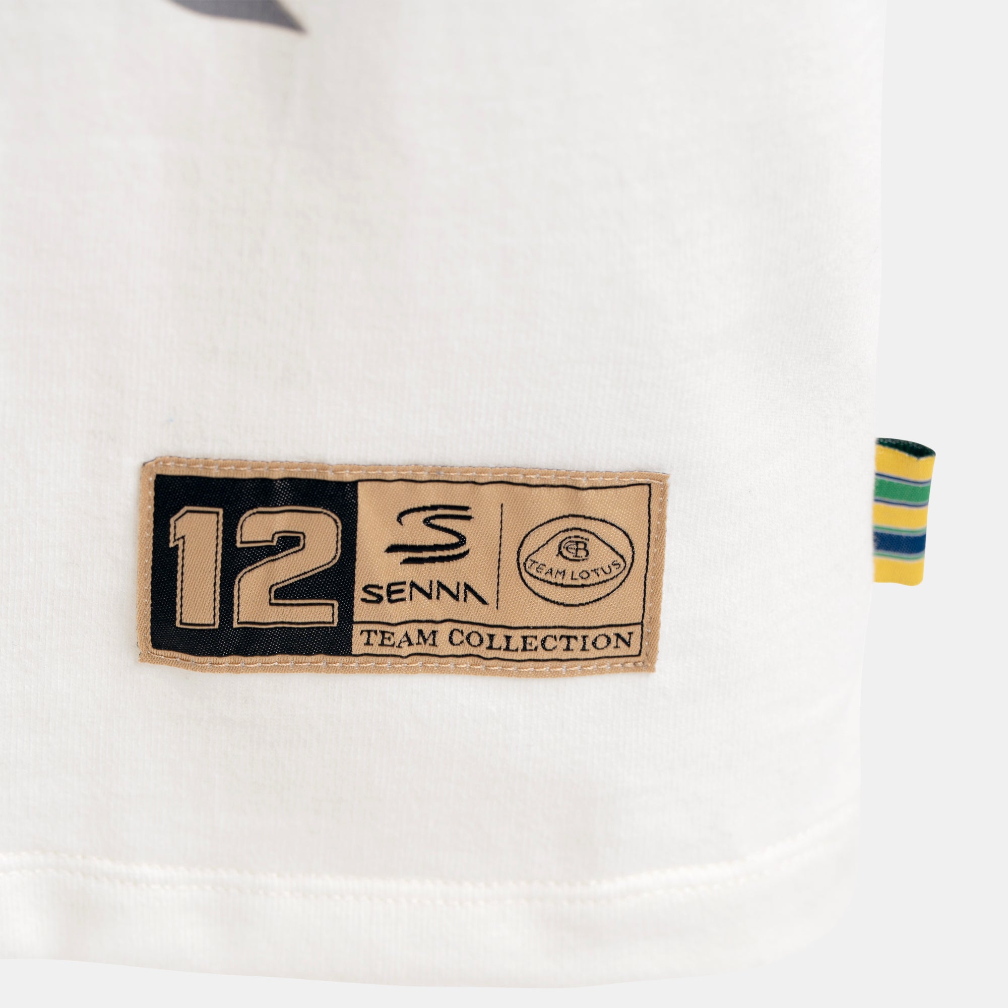 Close-up of a cream T-shirt corner showing a sewn-on patch with the number 12, Senna branding, and Team Lotus plus a small striped woven foldover tab on the right.
