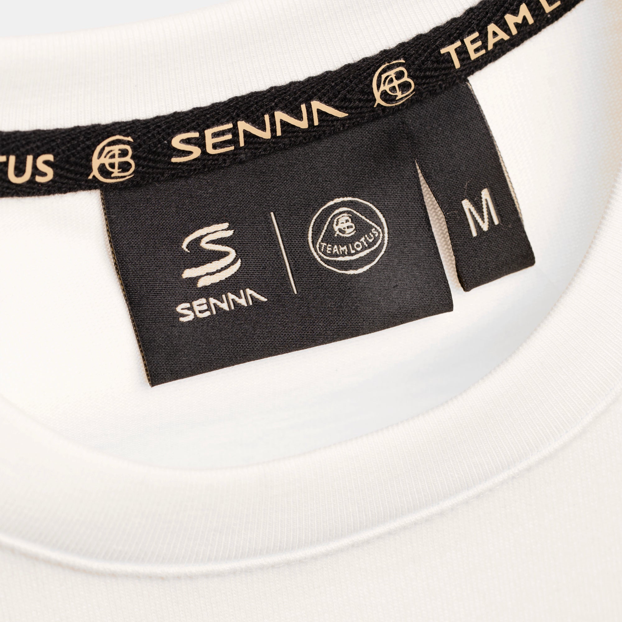 Close-up of a cream t-shirt showing the inner neck label and trim. The black neckband and labels are printed with the brand name SENNA and Team Lotus in pale gold, and the size tag reads M.