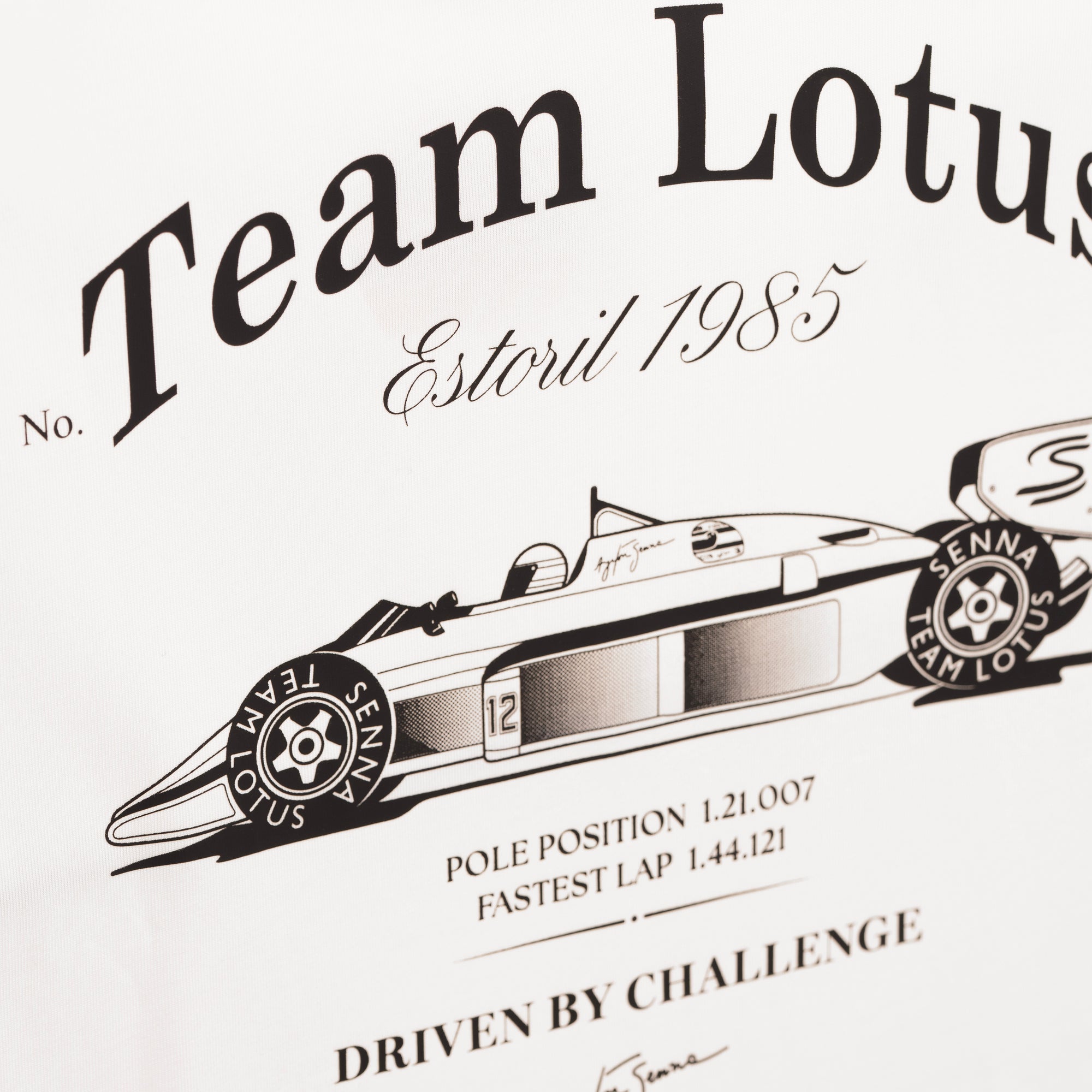 A close-up back side of a cream t-shirt printed with black vintage-style text reading 'Team Lotus Estoril 1985' and an illustration of a race car with 'Senna' tires and 'Team Lotus' branding.