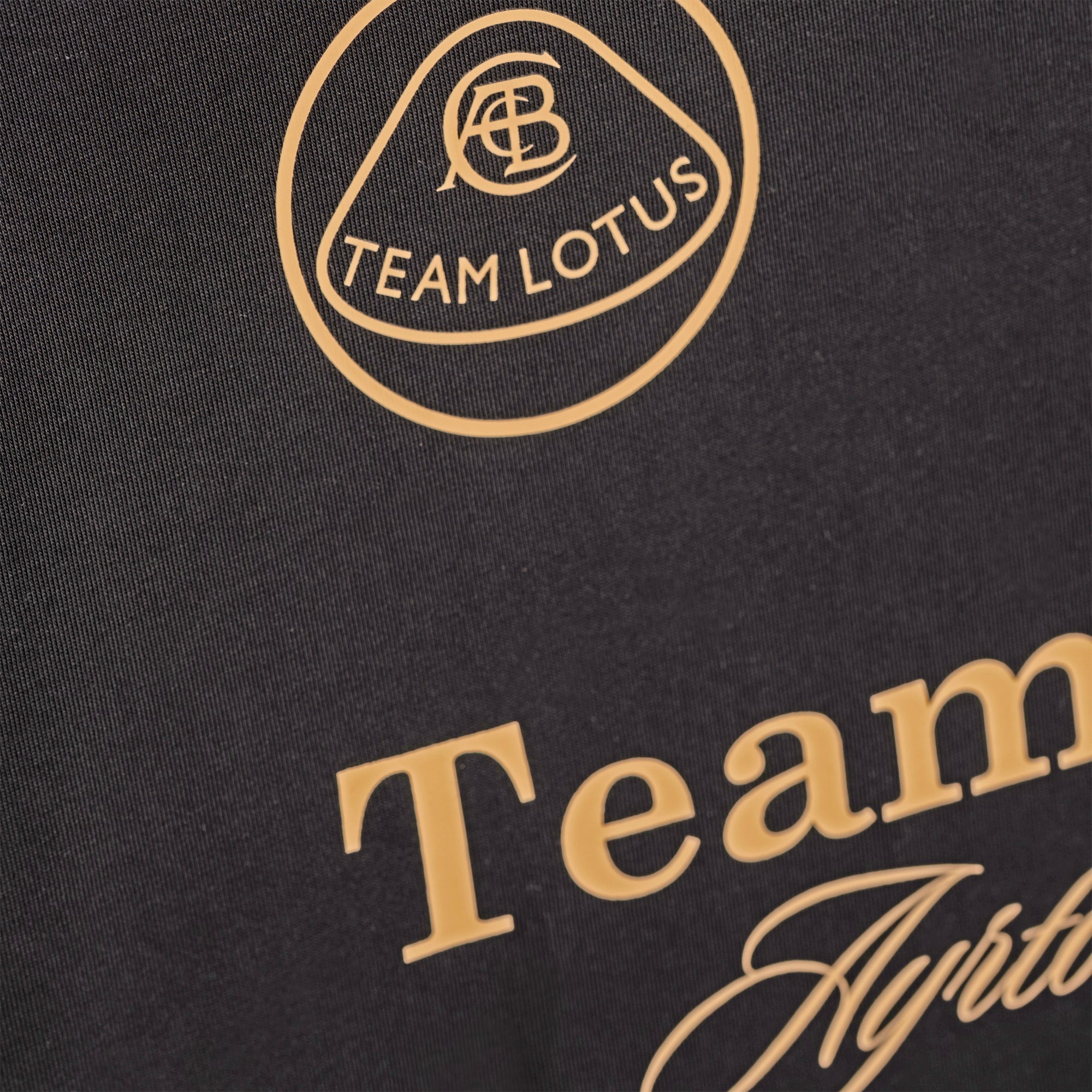 Close-up of a black oversize t-shirt with a gold circular Team Lotus logo and partial gold lettering of Team Lotus and script Ayrton Senna.