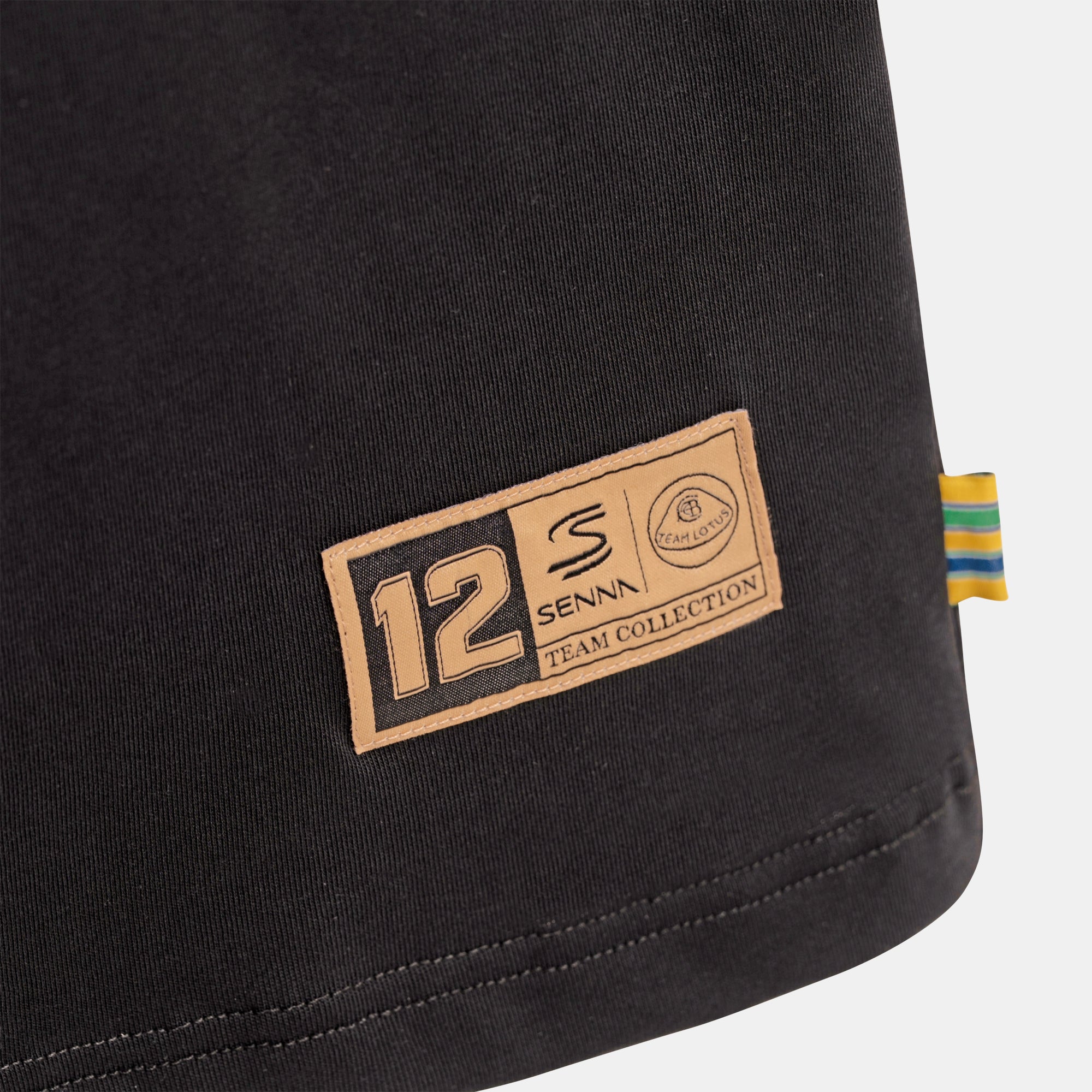Close-up of a black oversize tee showing a gold rectangular patch with the text '12 SENNA X TEAM LOTUS COLLECTION' and a small striped woven foldover tab on the right edge.