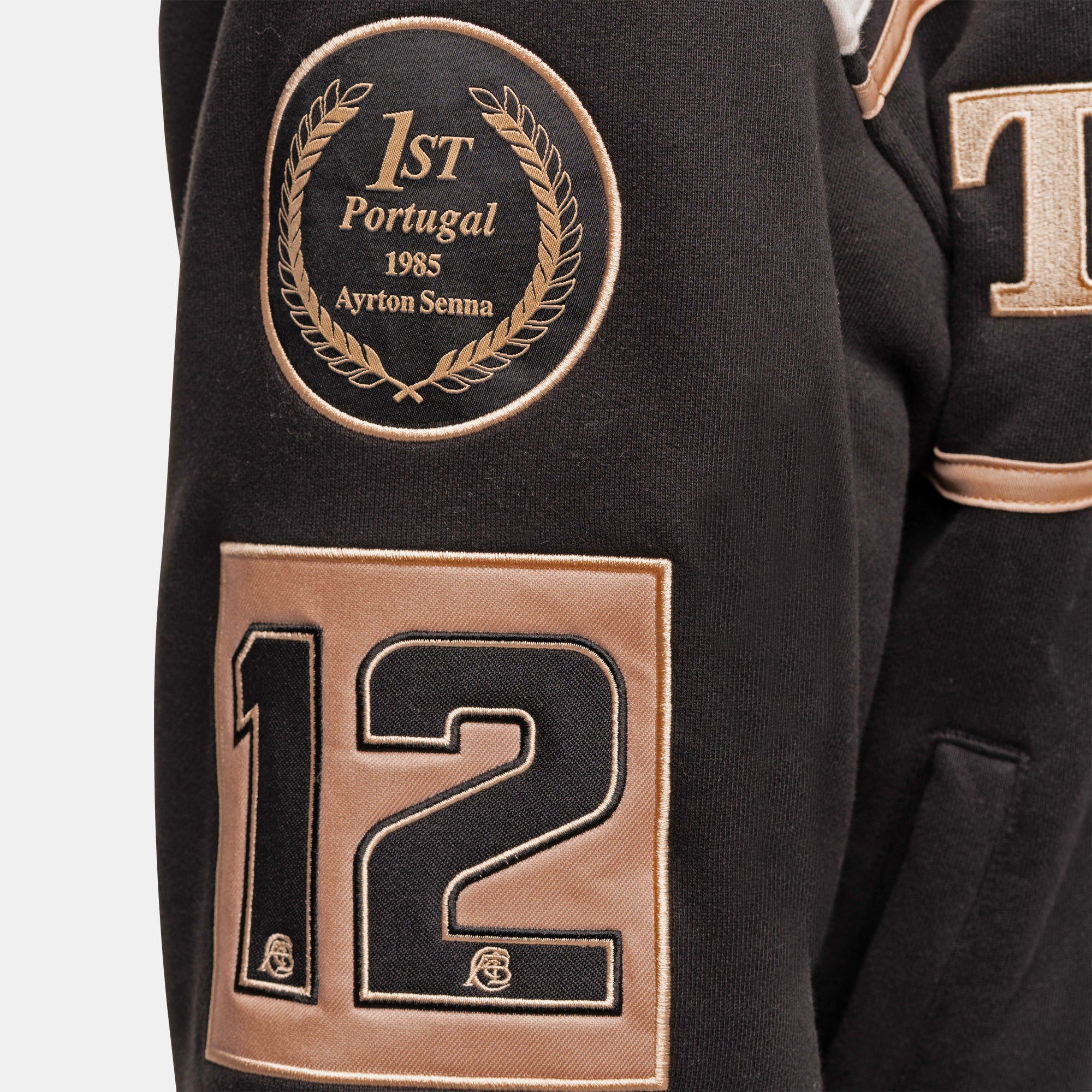 Close-up of a black embroidered jacket sleeve featuring a circular gold wreath patch reading '1st Portugal 1985 Ayrton Senna' and a large gold-and-black number 12 patch.