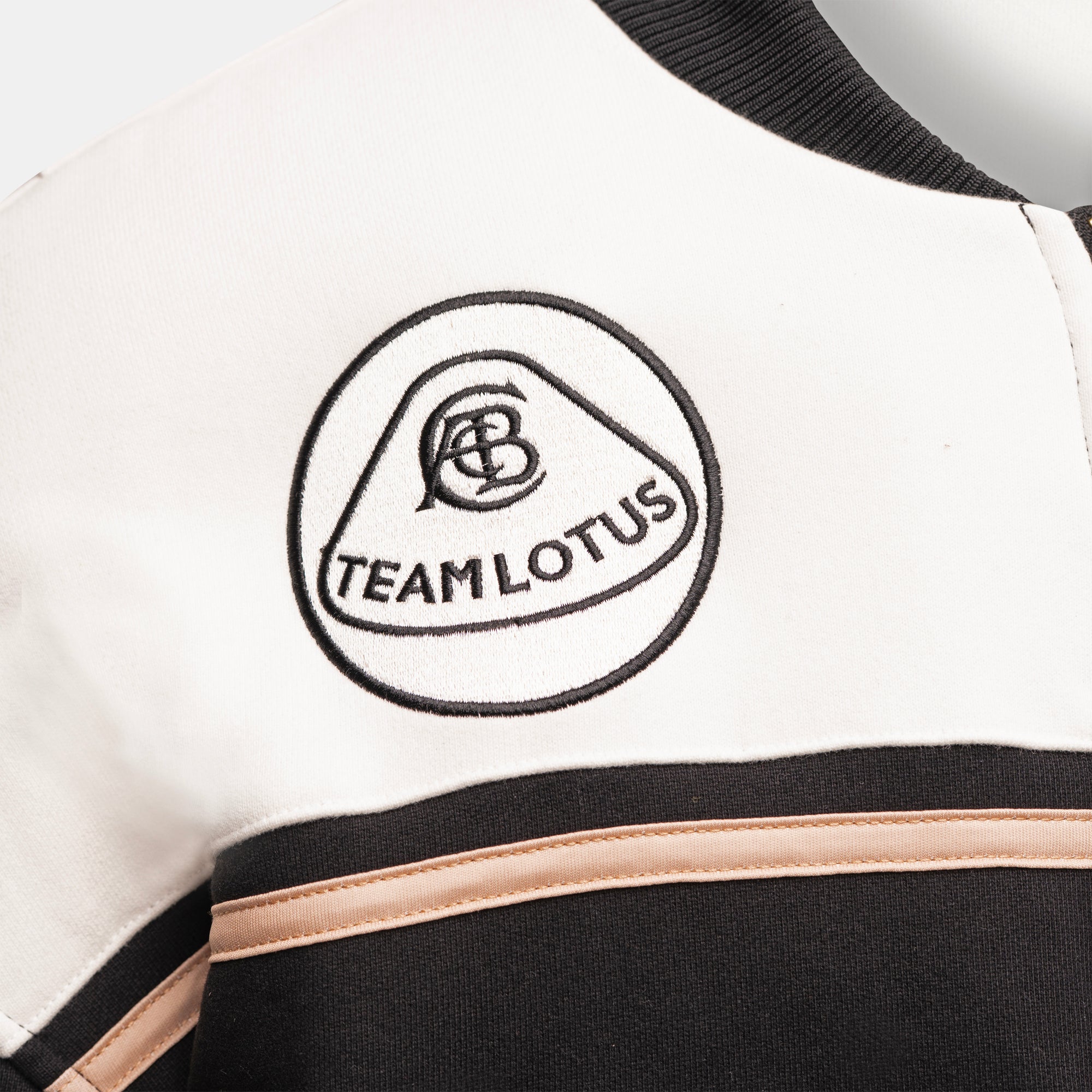Close-up of a Team Lotus embroidered logo on the chest of a white, black, and gold varsity-style jacket.