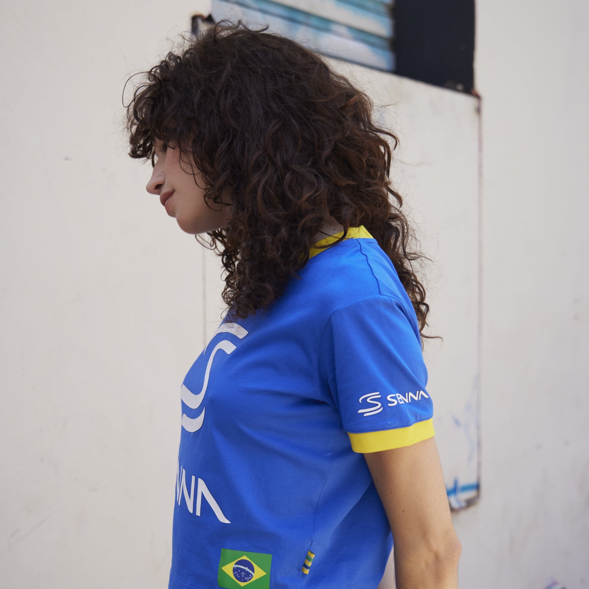 Person wearing a blue cropped tee with visible aryton senna branding, standing against a light-colored wall.