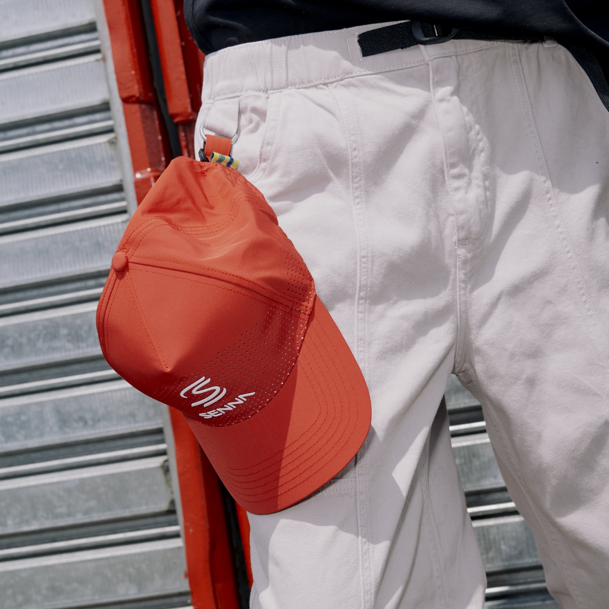Red Senna Leisure cap with a white logo held by a person wearing white pants against a metallic background