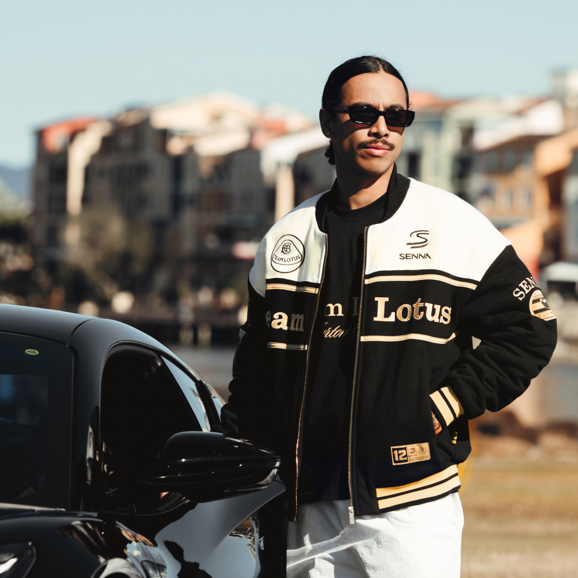 Male model wearing a black and white Senna Team Lotus varsity jacket beside a black sports car.