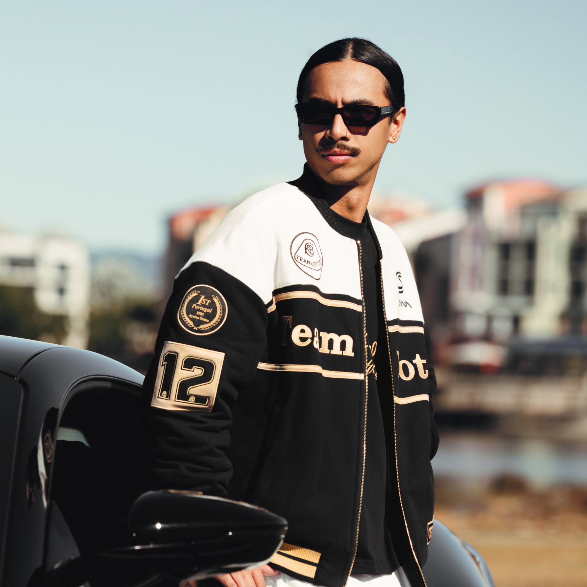 Male model wearing a Senna Team Lotus varsity jacket, showing the embroidered racing patches on the sleeve.