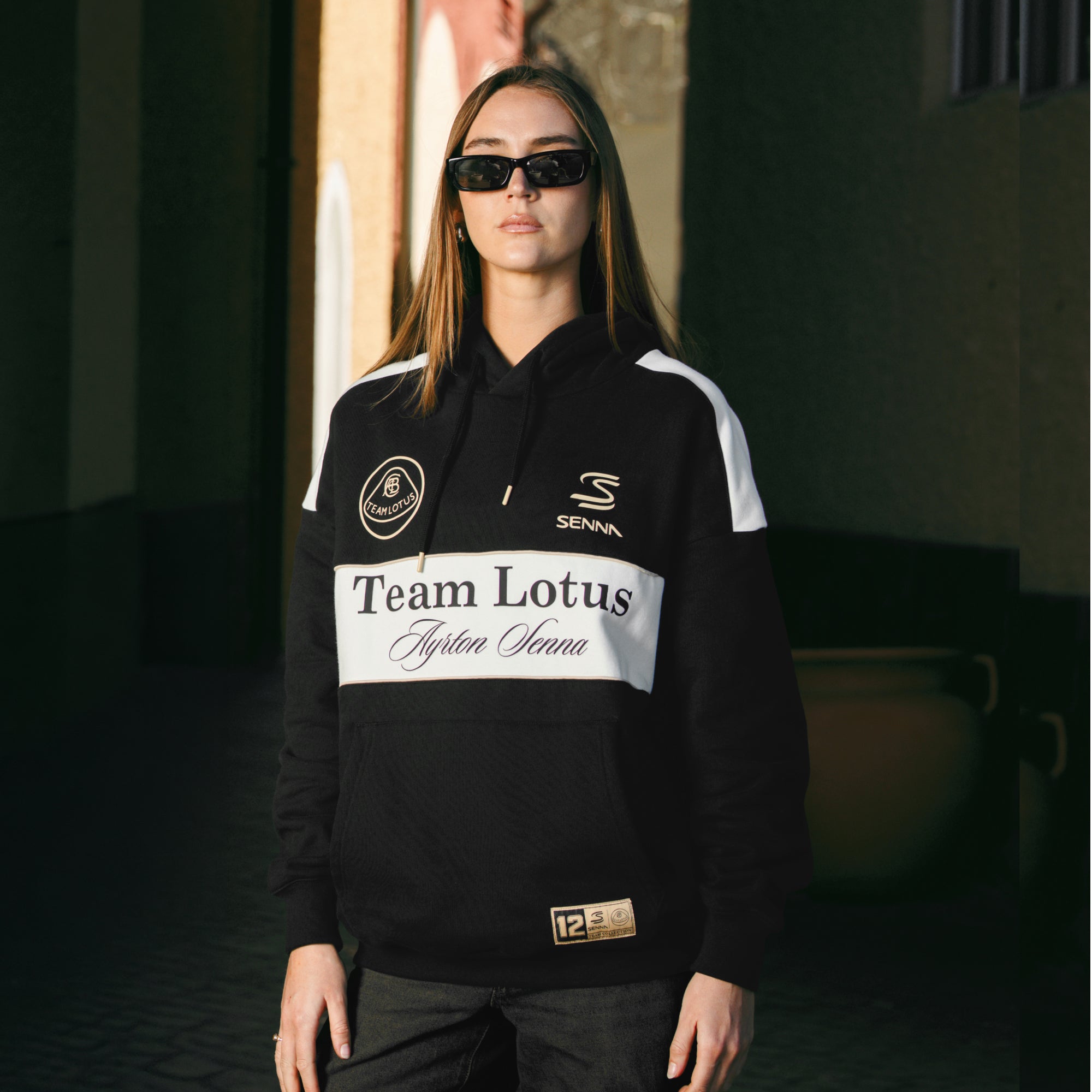 Female model wearing sunglasses and a black Team Lotus Ayrton Senna pullover hoodie.