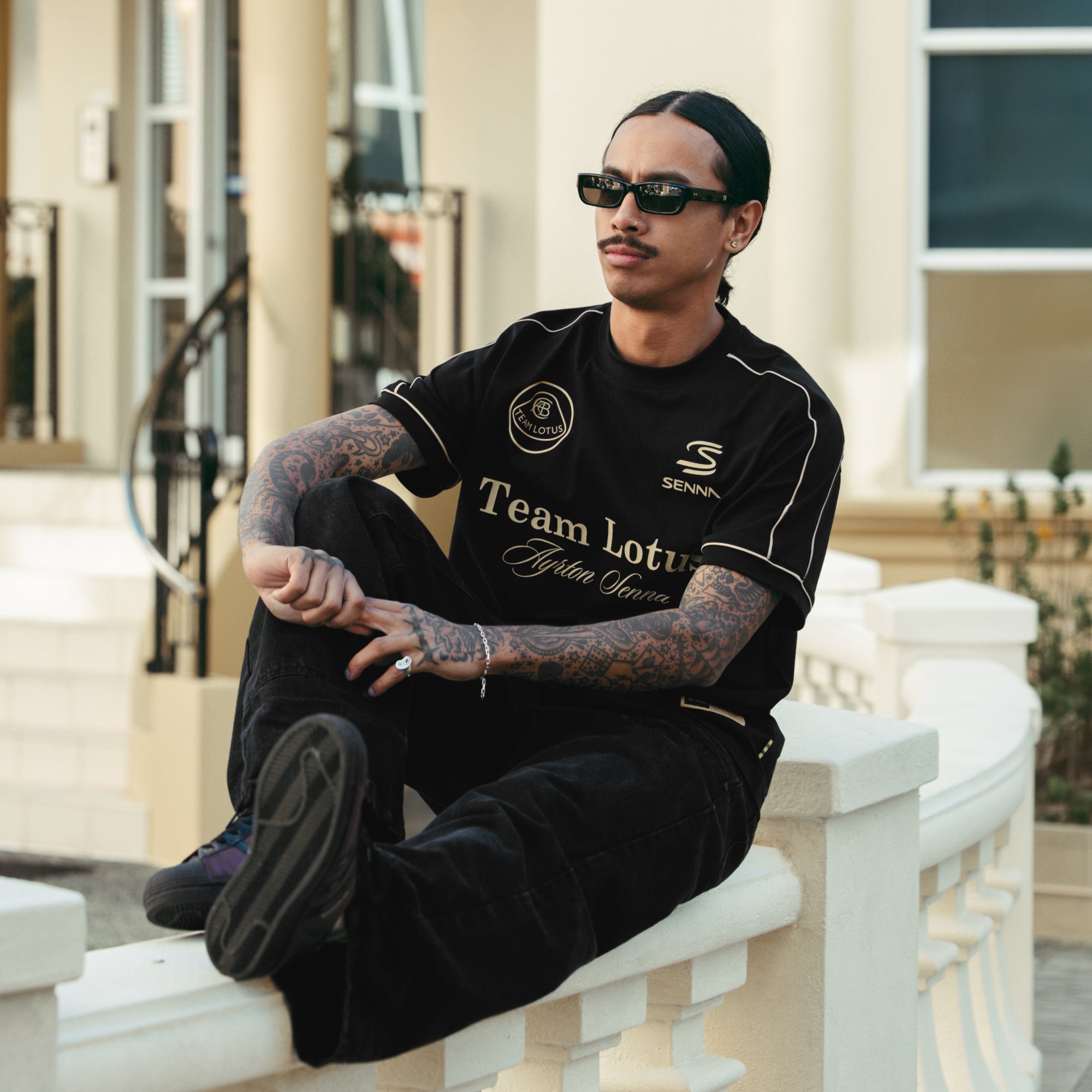 Man sitting on railing wearing a Team Lotus and Senna branded black and gold t-shirt.