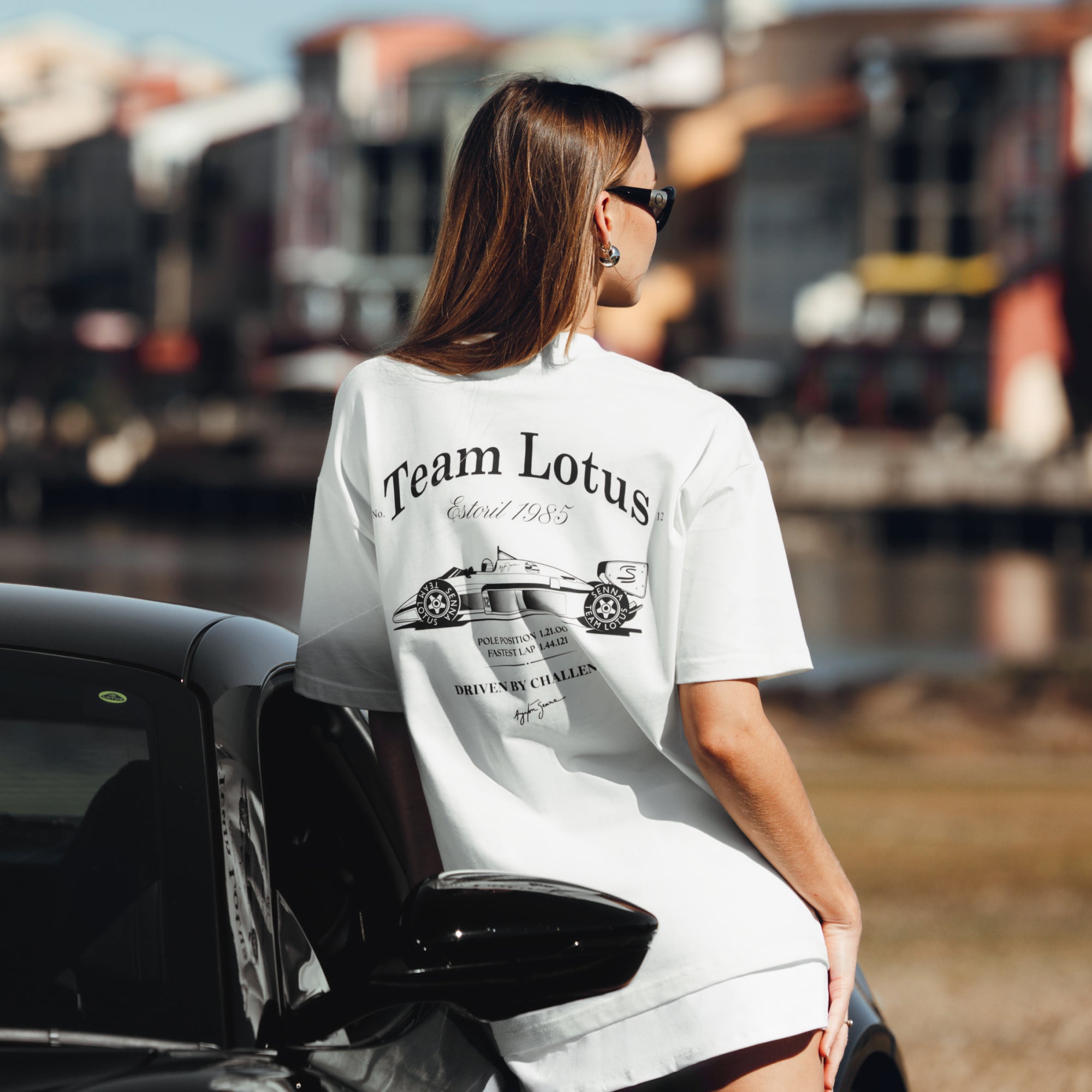 Woman leaning against car facing away while wearing a oversized cream tee with large Team Lotus graphic on back and Senna signature.