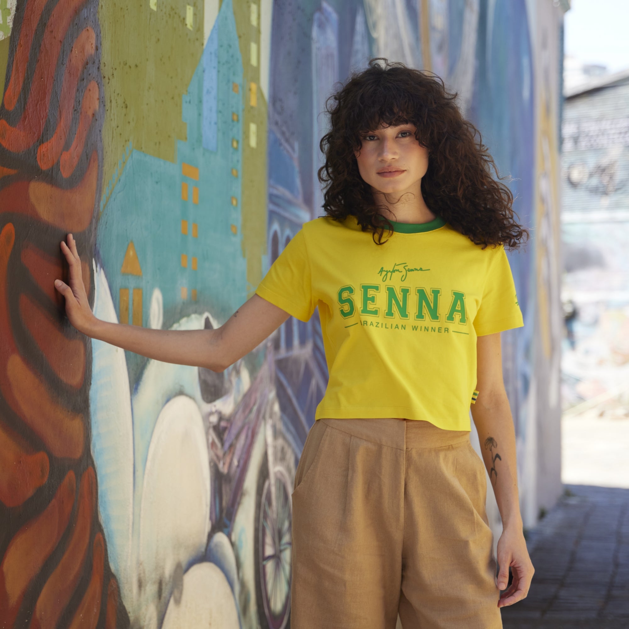 Woman wearing a yellow 'Senna' t-shirt in front of a colorful mural.