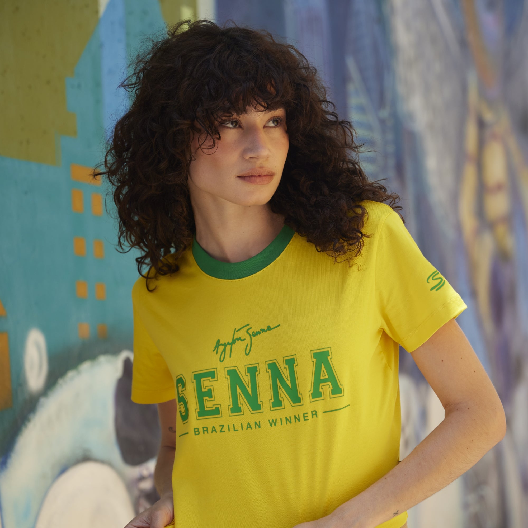 Woman wearing a yellow t-shirt with green text in front of a colorful mural.
