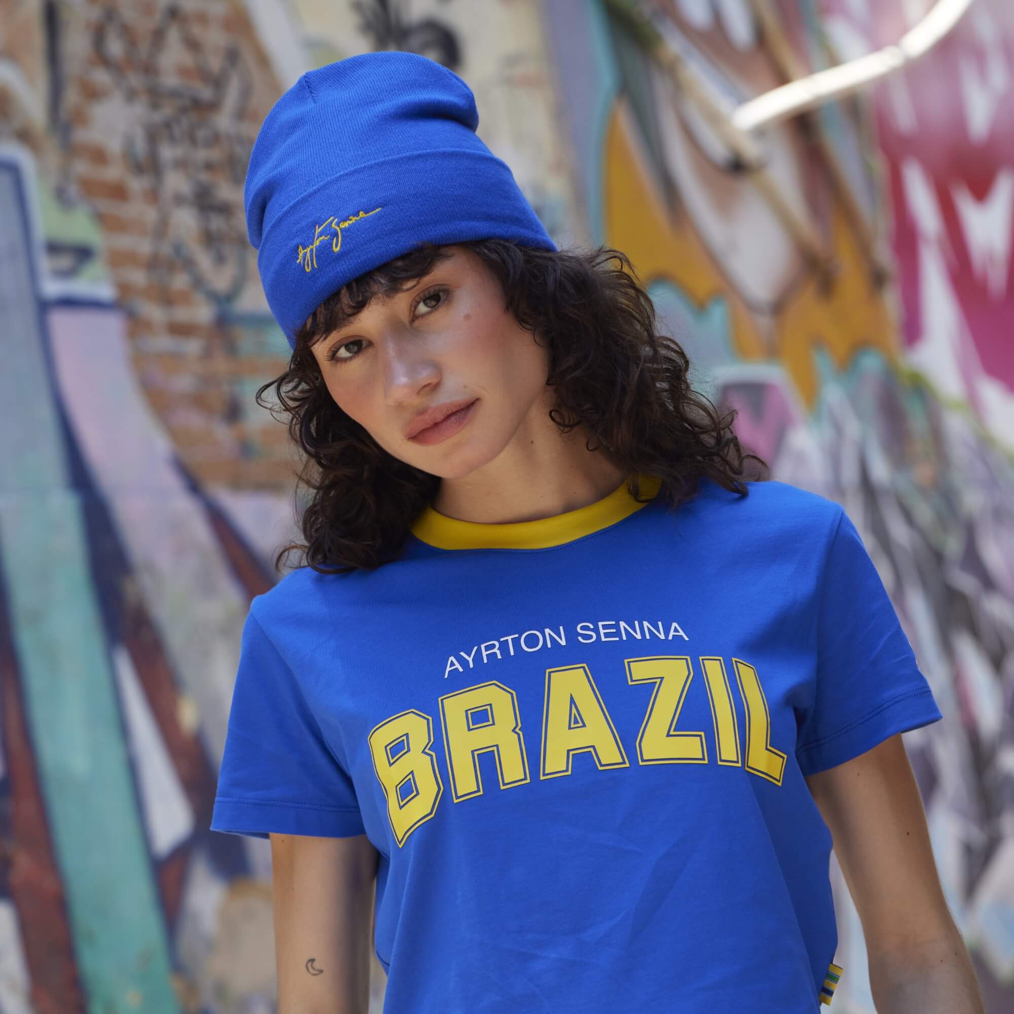 Person wearing a blue 'Ayrton Senna Brazil' t-shirt and blue beanie in front of a graffiti wall.