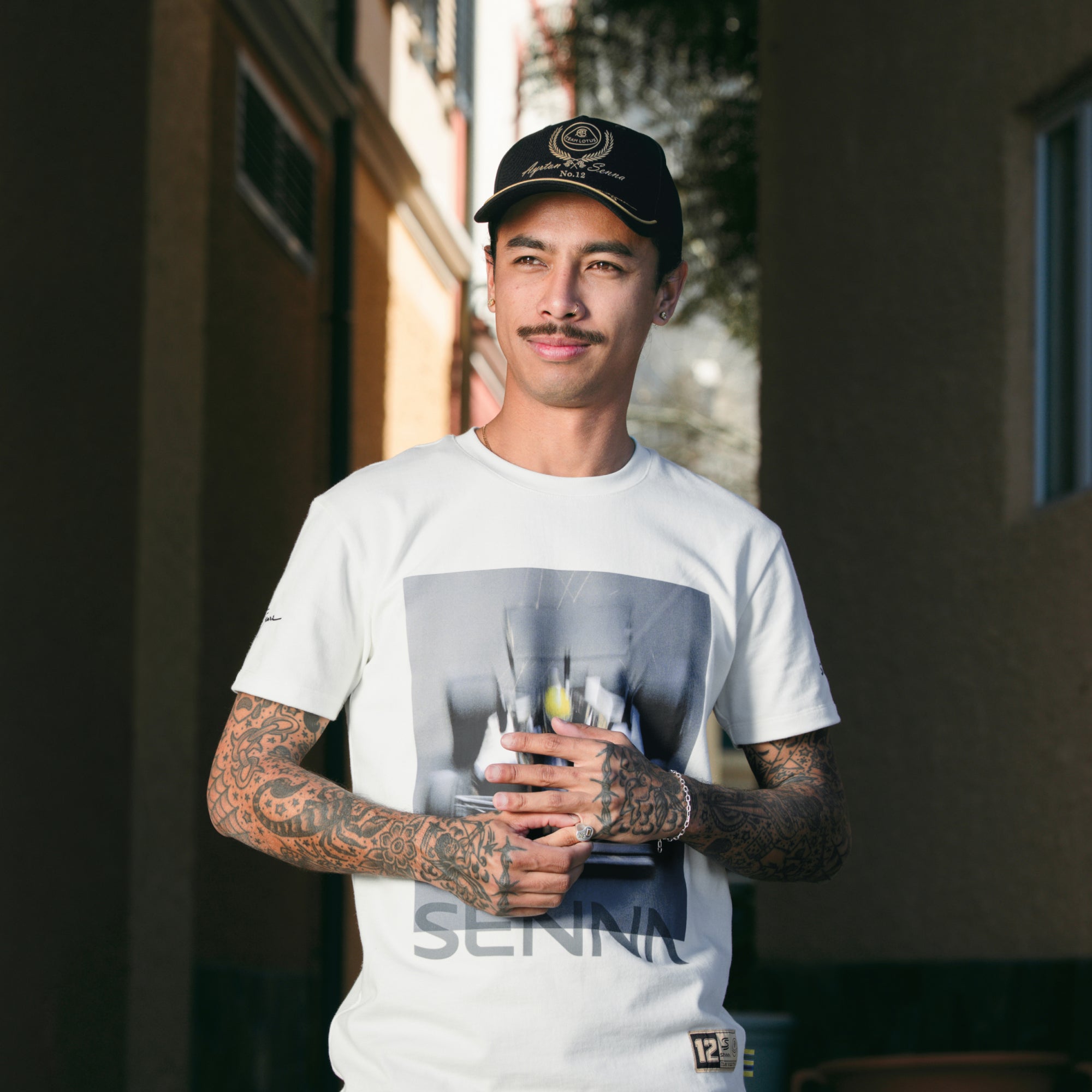 Cream t-shirt with large Senna type and race graphic worn by casual posed man in alley way.