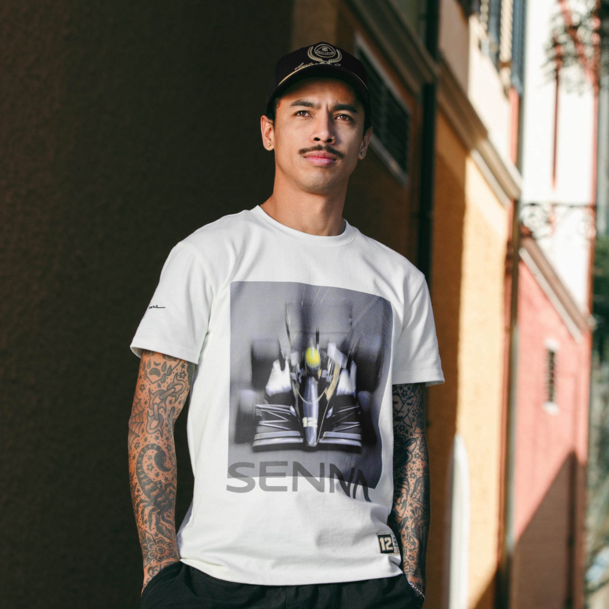 Man standing in alley wearing a cream tee with Senna Formula 1 graphic print on front and Team Lotus collaboration.
