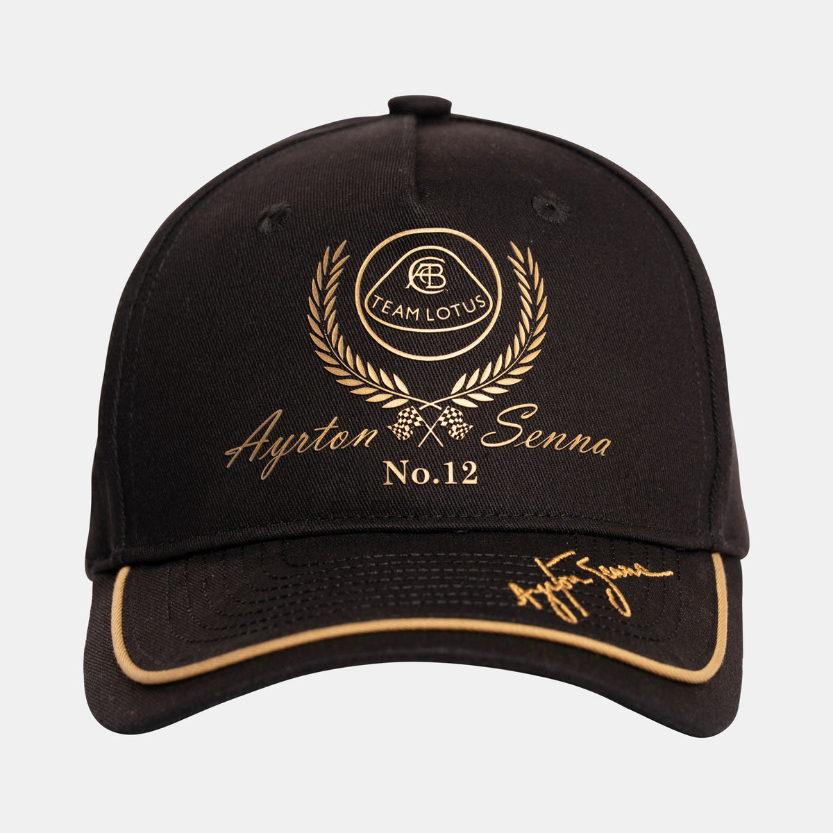 A black Team lotus and Senna race cap with gold embroidered details, featuring a 'Team Lotus' laurel wreath crest, 'Ayrton Senna No.12' text, and a signature on the brim.