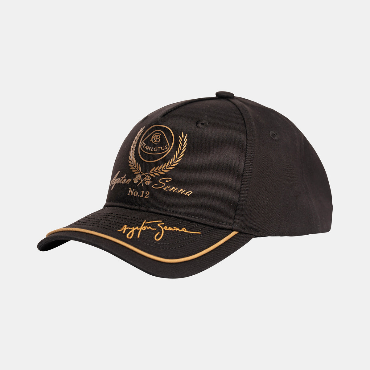A black race cap with gold embroidered details, featuring a crest with a laurel wreath, and the text 'Team Lotus Ayrton Senna No.12' along with a stylized signature on the brim, set against a light gray background.