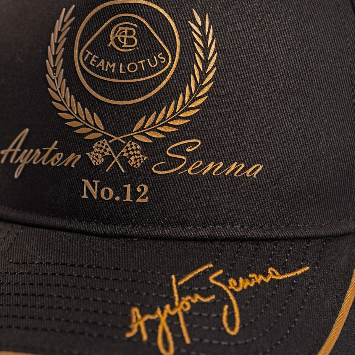 Close-up of a black Team Lotus and Senna race cap featuring gold embroidered details, including a Team Lotus laurel wreath crest, 'Ayrton Senna No.12' text, and a gold signature on the brim.