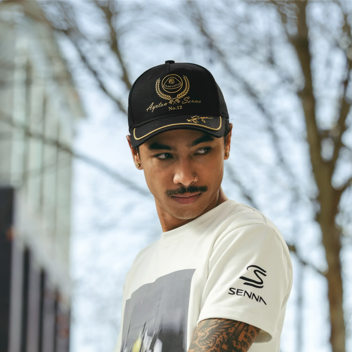 A man wearing a black and gold Team Lotus and Senna Racing cap and a white Senna t-shirt, photographed outdoors against a blurred background of trees and a building.